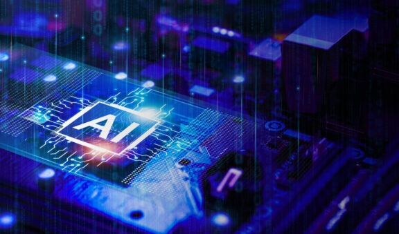 Close-up of a computer processor with a glowing ‘AI’ symbol on the chip, representing artificial intelligence technology. The circuit board and surrounding components are illuminated in blue and purple tones, giving a futuristic and high-tech appearance.